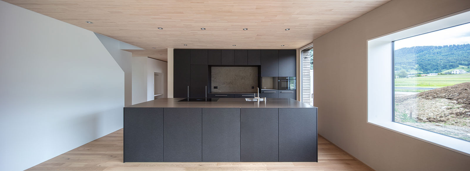 Blick auf die zweifronten Küche aus schwarzem MDF mit Chromstahlabdeckung. Die schwarze Küche ist im Kontrast du den weissen Wänden. Decke und Boden in hellem Holz.