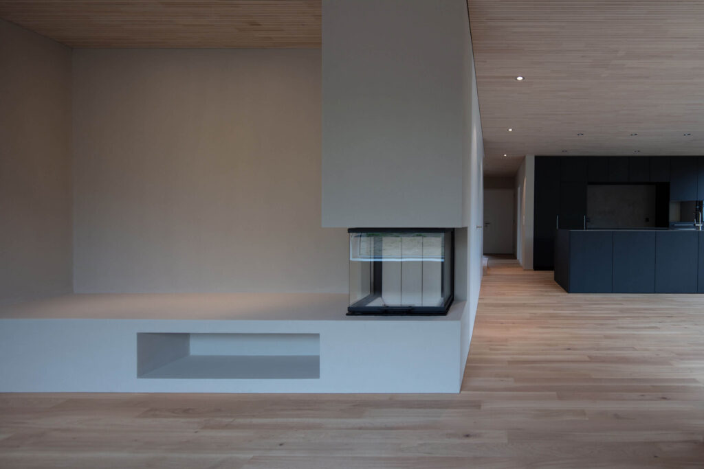 Eine Wand mit Sitzbank und integriertem Cheminée im Wohnbereich. Im Hintergrund die Küche aus schwarzem MDF.