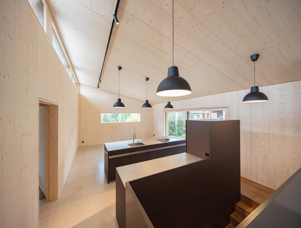 Der Wohnraum ist schlicht gehalten und materialisiert: Die Wände und das Dach sind in Holz Natur belassen, dunkle Einbaumöbel und Beleuchtungskörper kontrastieren das Holz.