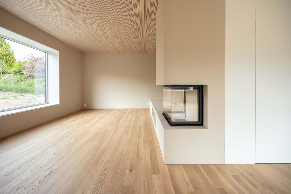 Die Holzlammellendecke, der sandige Farbton der Wände und der Holzboden erzeugen eine warme Stimmung im Wohnbereich mit Cheminée. Ein grosszügiges Fenster nimmt Bezug ins Freie.