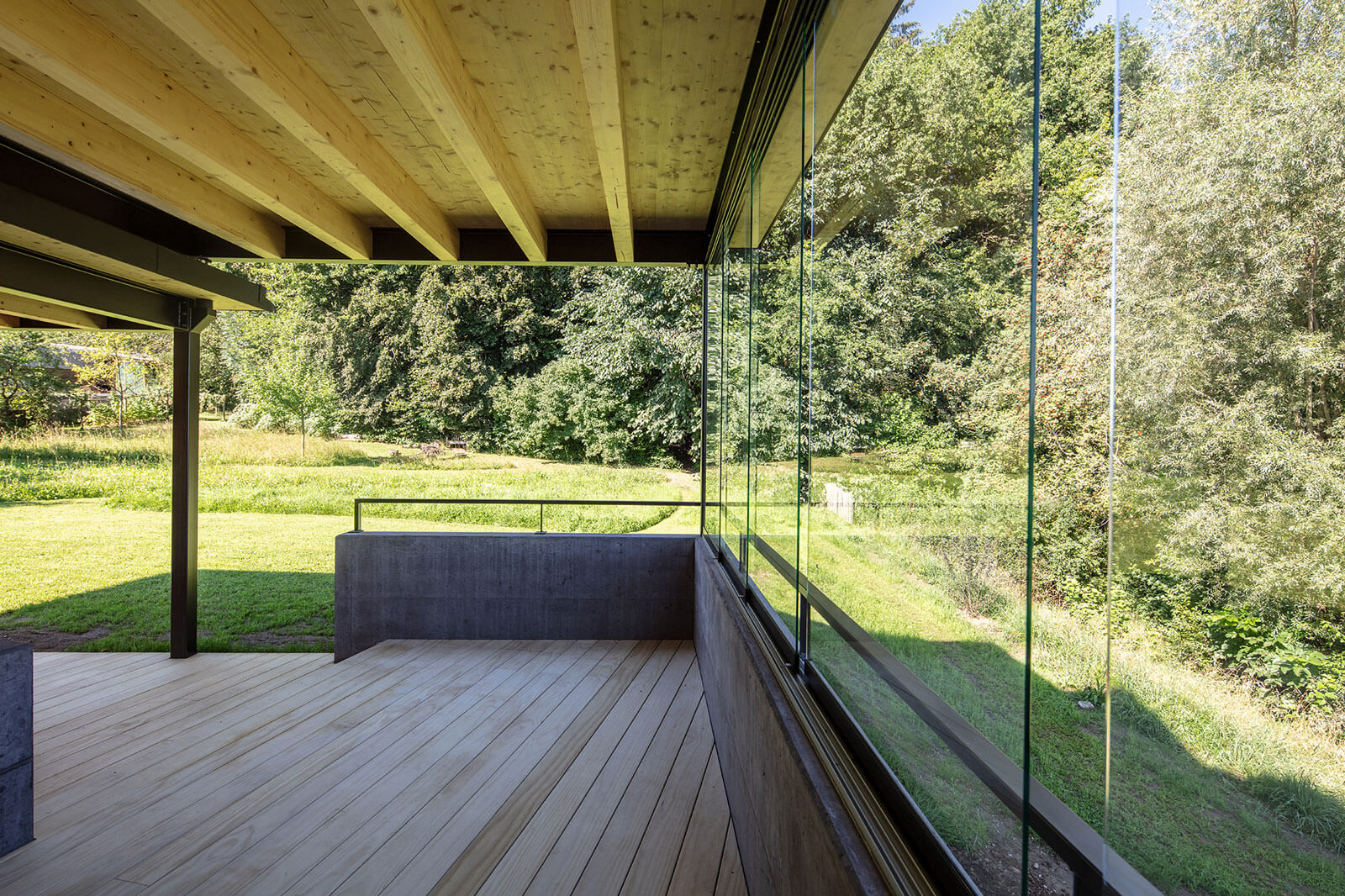 Blick vom Sitzplatz auf den Waldrand. Schwarz eingefärbter Beton, schwarzer Stahl in Kombination mit Holz bieten der Familie eine Erweiterung des bestehenden Wohnraumes.
