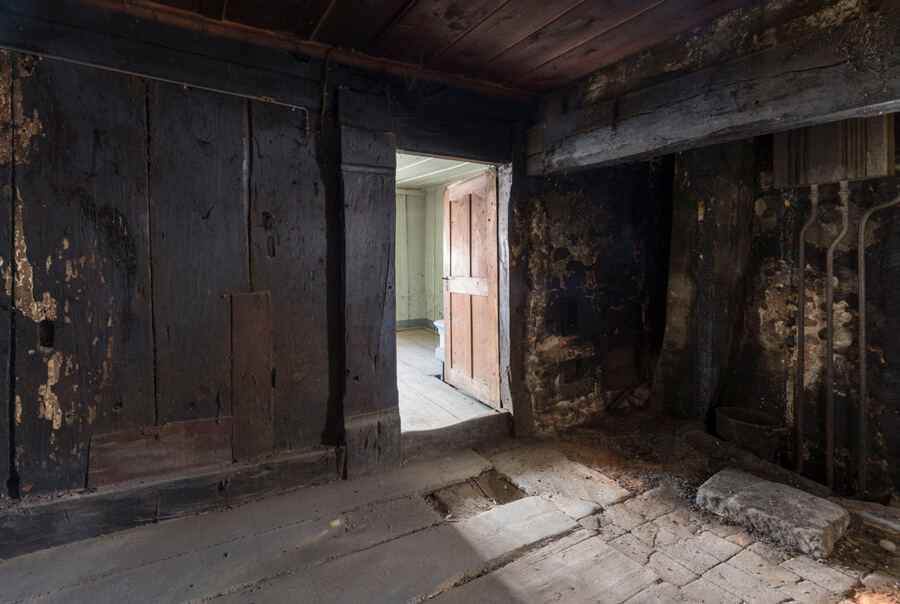 Von einem dunklen Raum mit geschwärzten Holzbalken gelangt man durch eine niedrige Türe über eine Stufen in einen hellen, vertäferten und lindgrün gestrichenen Raum.