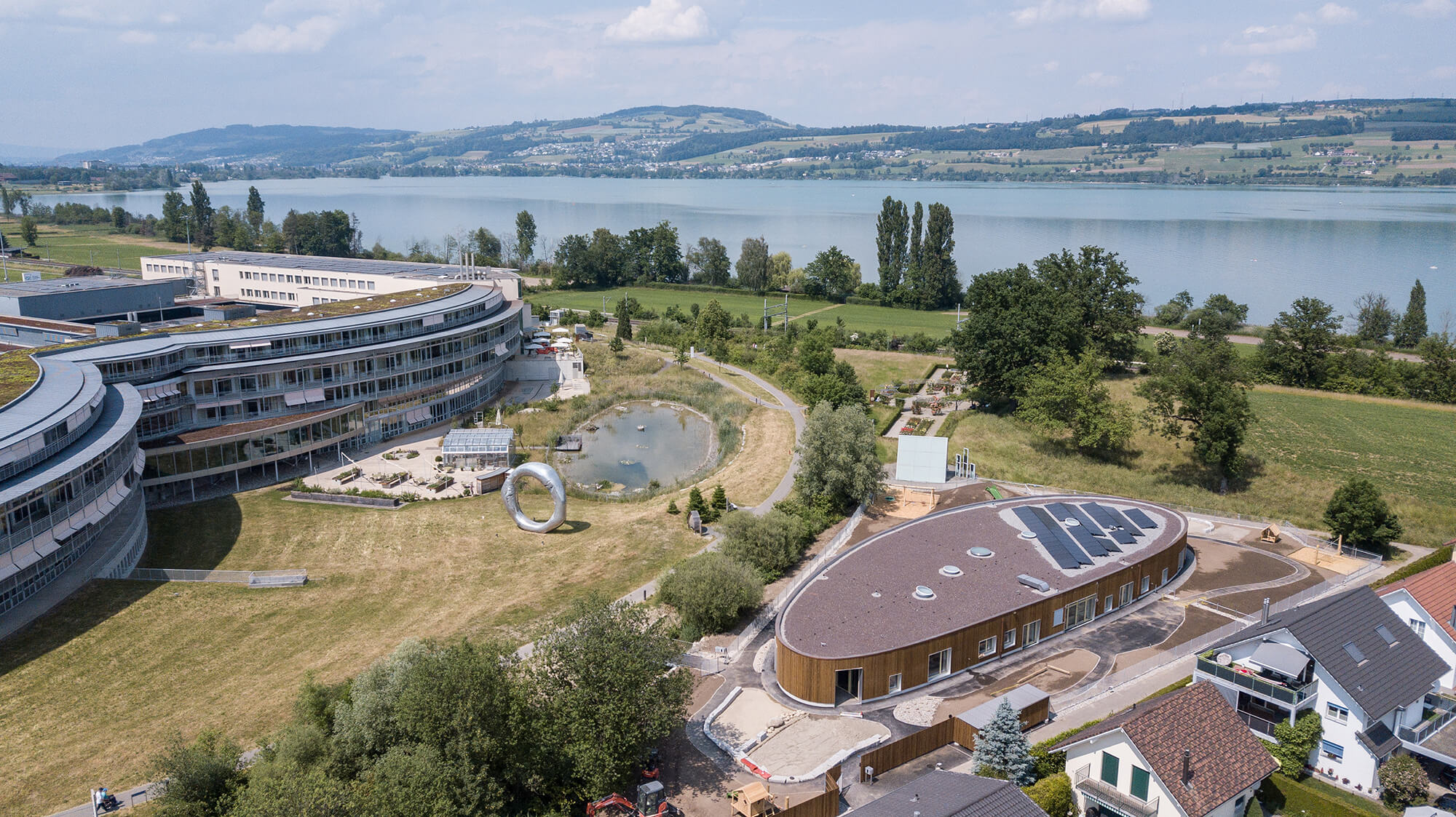 Vogelperspektive auf das Gelände vom Schweizer Paraplegiker Zentrum mit dem Neubau der Kita, dem ovalen Teil, dem gerundeten Spitaltrakt und dem Sempachersee im Hintergrund.