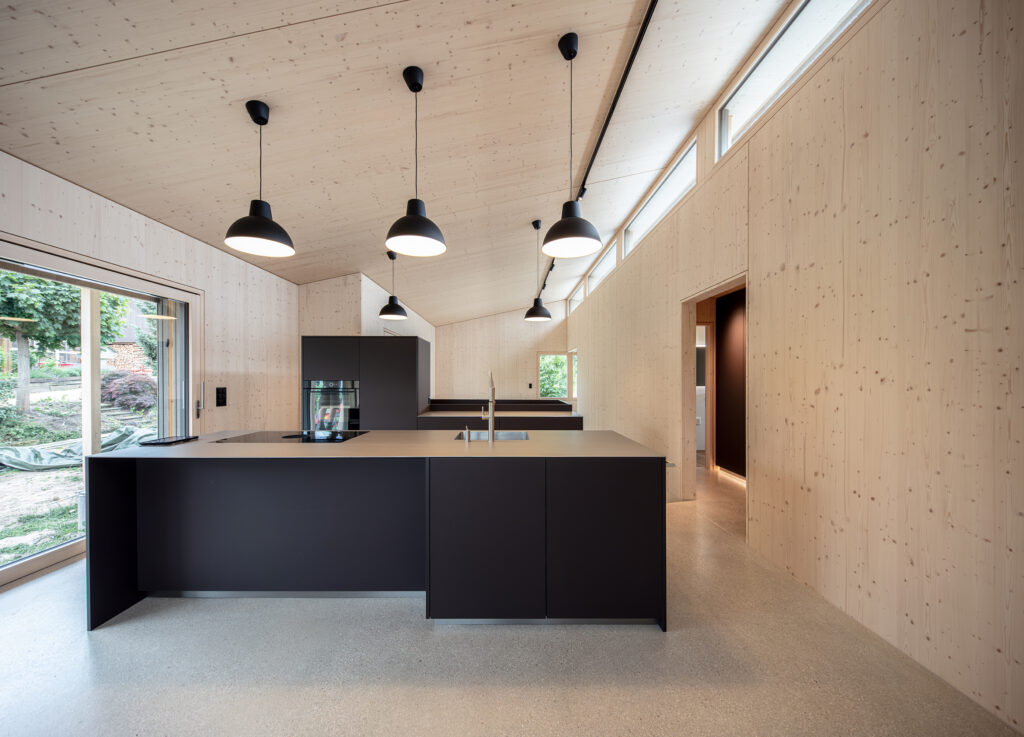 Eine offene Küche mit Ess- und Wohnbereich unter einem Schrägdach. Wände und Decke sind aus Tannenholz natur.